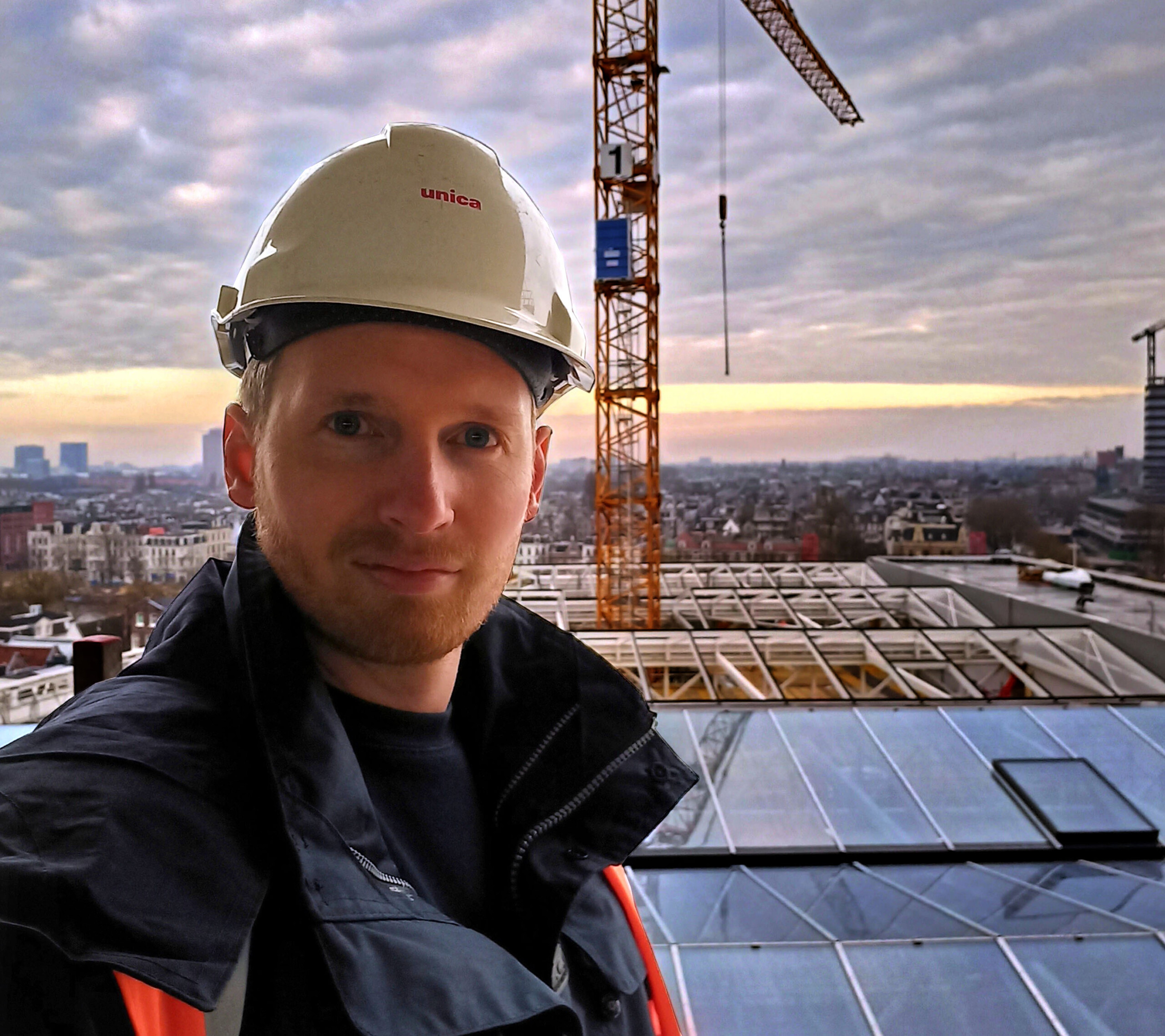 Rogier Koops kijkt in de camera. Hij staat op het dak van een gebouw met op de achtergrond een hijskraan en een ondergaande zon.